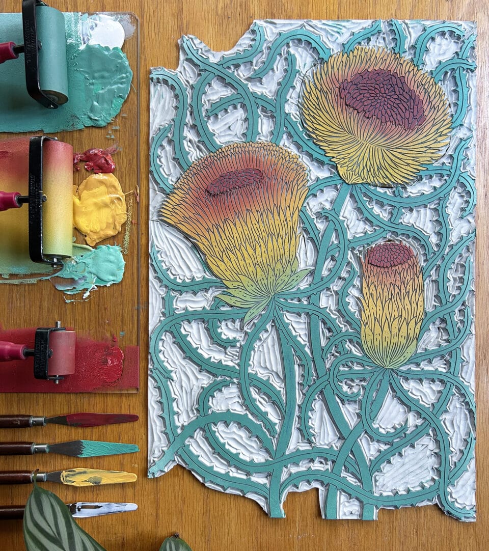 a printing block with a carving of three thistle-like flowers, laid on a wooden table with inks and tools set next to it