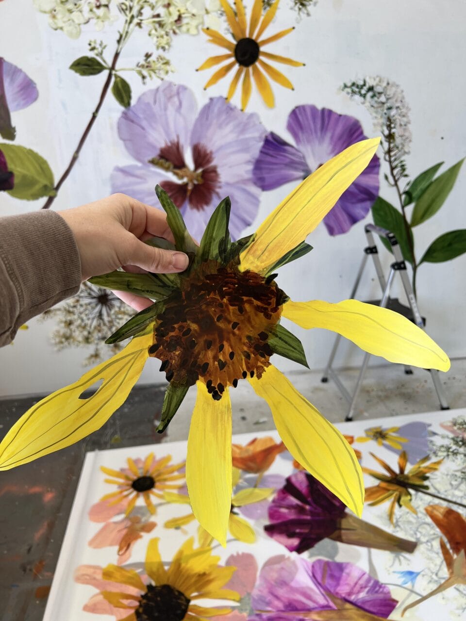 a hand holds a large painted cutout of a flower, in front of numerous others already installed on a wall, to show a realistic rendering of a large-scale yellow bloom that looks pressed